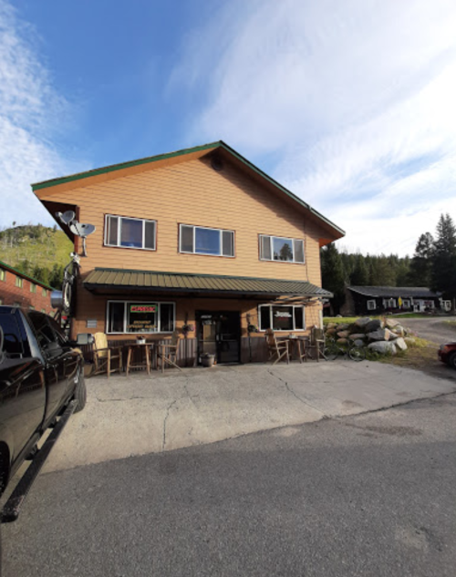 A two-story wooden building with a green roof, featuring a porch and outdoor seating, surrounded by trees and mountains.