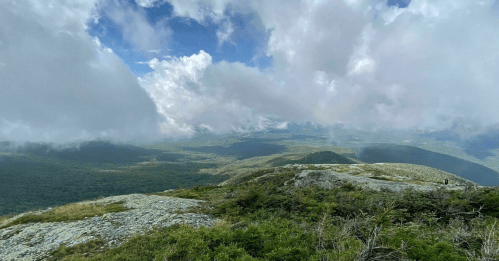 A panoramic view from a mountain summit, featuring lush green valleys and dramatic clouds in a blue sky.