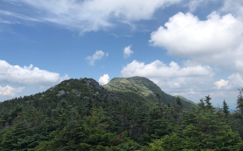 A scenic view of two mountains surrounded by lush green trees under a partly cloudy blue sky.