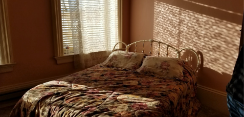 A sunlit bedroom with a floral bedspread, casting patterned shadows on the wall from the window.