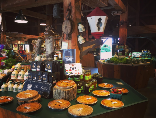 A rustic market display featuring pies, coffee, jams, and fresh produce in a wooden barn setting.