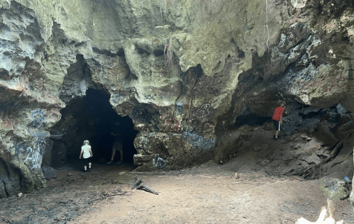 Two children explore a rocky cave with graffiti on the walls, surrounded by lush greenery.
