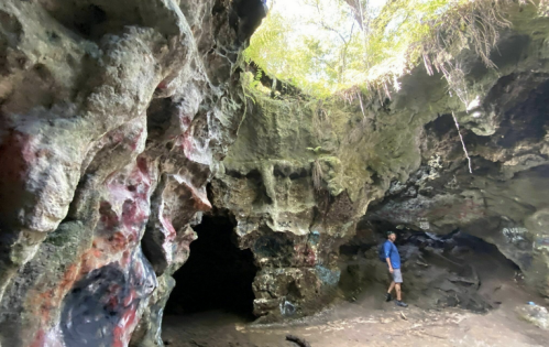 A person stands inside a rocky cave surrounded by colorful rock formations and lush greenery.