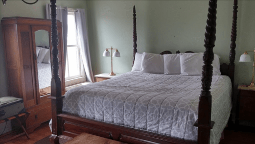 A cozy bedroom featuring a four-poster bed, two lamps, and a vintage wardrobe, with natural light from a window.
