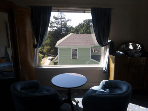 A cozy room with two blue chairs and a small table, featuring a large window overlooking a green house and trees.