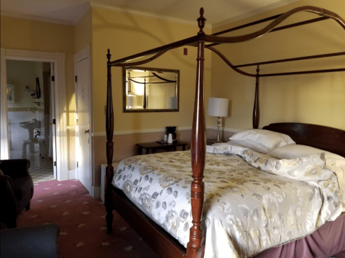 Cozy bedroom with a four-poster bed, soft lighting, and a mirror, featuring a seating area and an adjoining bathroom.