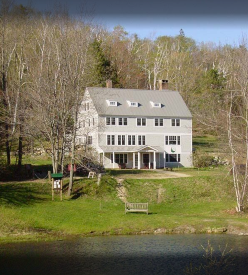 A large gray house with a porch, surrounded by trees and a pond, set in a serene natural landscape.
