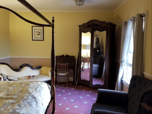 Cozy bedroom featuring a four-poster bed, vintage mirror, armchair, and soft lighting.