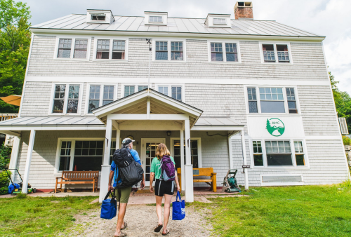 Two hikers approach a rustic lodge with backpacks, ready for an outdoor adventure. Green trees surround the building.