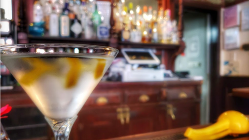 A close-up of a martini glass with lemon twists, set against a blurred bar with bottles in the background.