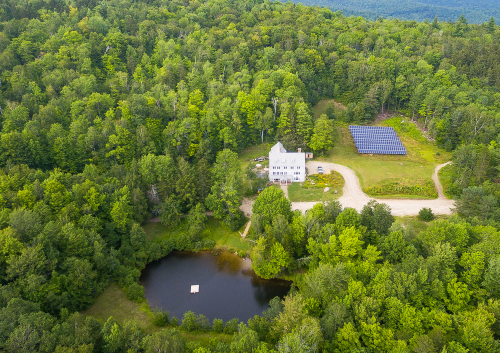 Aerial view of a house near a pond, surrounded by lush green trees and a solar panel array in the background.