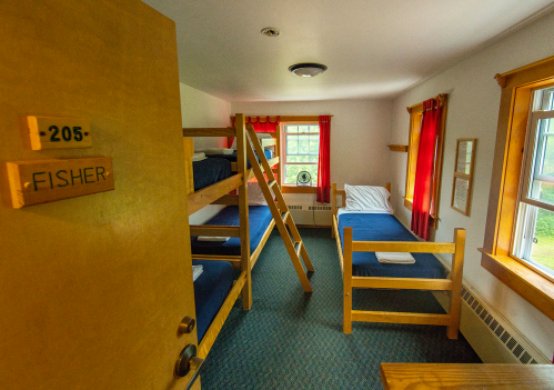 A cozy dorm room with two bunk beds, blue bedding, and red curtains, featuring a door labeled "Fisher" and a window.