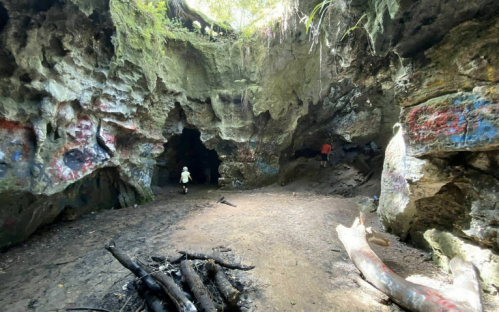 A rocky cave interior with colorful graffiti, featuring two people exploring and natural light filtering in.