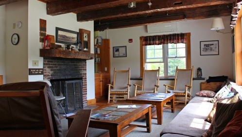 Cozy living room with wooden beams, a fireplace, rocking chairs, and a sofa, featuring natural light from a window.