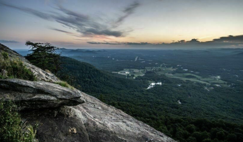 A panoramic view of a lush valley at sunset, with mountains in the distance and a rocky foreground.