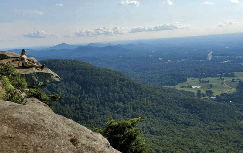 A person sits on a rocky ledge, overlooking a vast green valley and distant mountains under a clear blue sky.