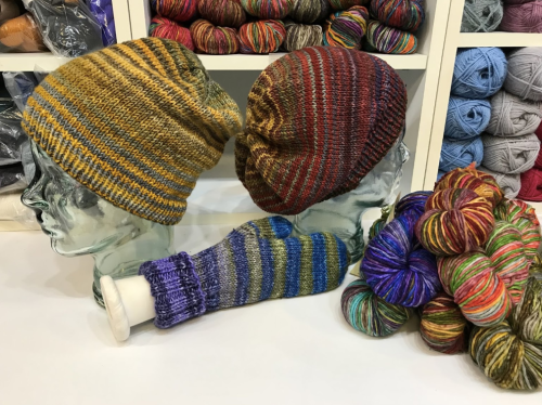 Two knitted hats displayed on glass heads, with a colorful striped arm warmer and a pile of vibrant yarn in the foreground.