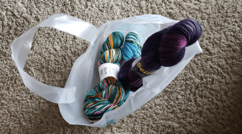 A white plastic bag containing colorful yarn skeins in blue, green, purple, and multicolored patterns on a textured surface.