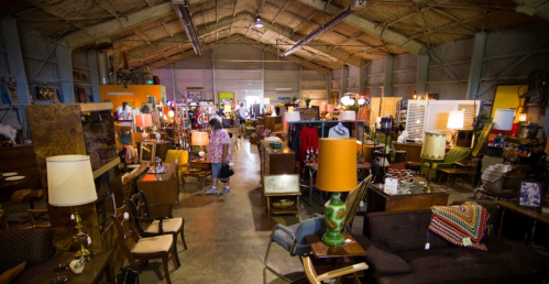 A spacious vintage store filled with various furniture, lamps, and decor items, with a person browsing in the aisle.