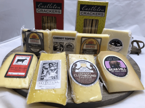 A wooden platter displays various cheeses and crackers, including labels from Vermont and Cave Aged varieties.