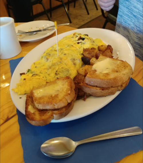 A plate of breakfast featuring an omelette, home fries, and French toast, with a spoon and coffee cup in the background.