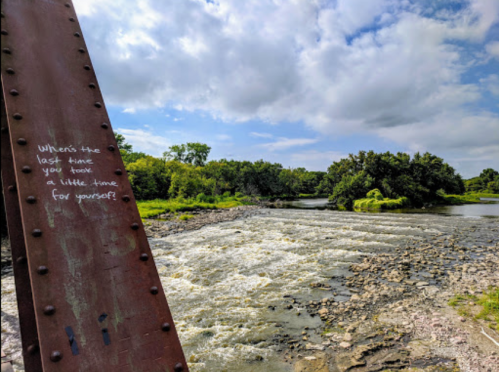 A river flows beside a rusted metal structure, with trees and clouds in the background. A message is written on the metal.