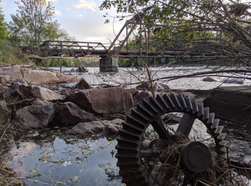 A rusted gear lies in the foreground by a river, with a dilapidated bridge visible in the background among trees.