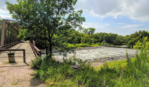 A scenic view of a river with rapids, framed by trees and a bridge in a lush, green landscape under a cloudy sky.