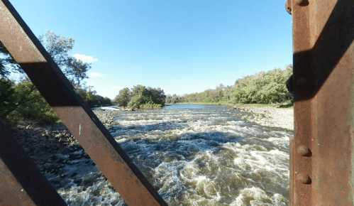 View of a river flowing through a lush landscape, framed by rusty metal beams under a clear blue sky.
