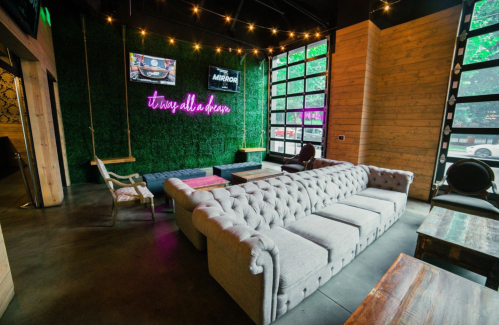 Cozy lounge area with a tufted gray sofa, greenery wall, neon sign, and large windows letting in natural light.