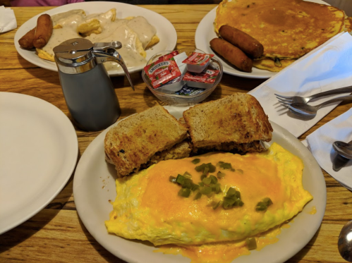 A plate with a cheese omelet topped with green peppers, toast, and a side of pancakes and sausages. Syrup and condiments included.
