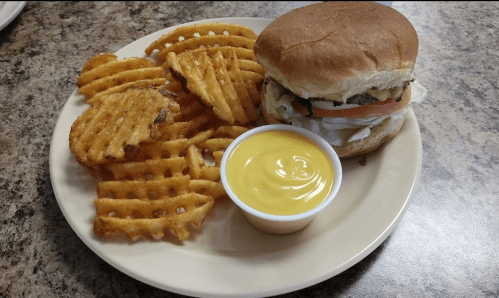 A plate with a burger topped with lettuce and tomato, served with waffle fries and a small cup of yellow dipping sauce.