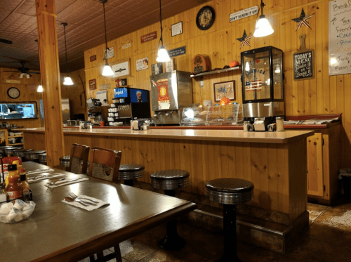 Cozy diner interior with wooden walls, a counter, stools, and tables set for dining, featuring condiments and decor.