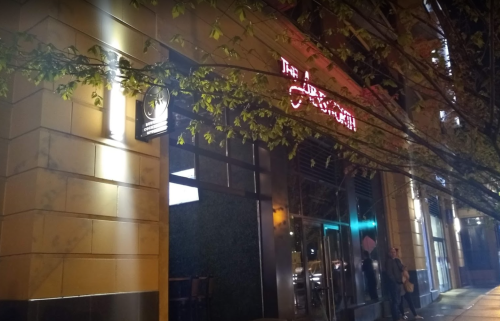 A dimly lit street view of a restaurant with neon signage, partially obscured by tree branches.