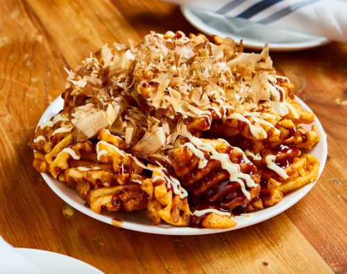 A plate of waffle fries topped with bonito flakes, drizzled with sauce and mayonnaise, on a wooden table.