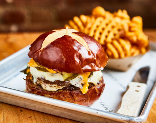 A juicy burger with cheese, pickles, and an egg on a pretzel bun, served with a side of waffle fries.