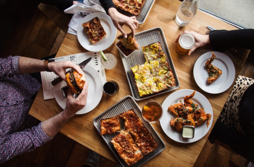 A table filled with various dishes, including pizza, fried chicken, and drinks, as people enjoy a meal together.