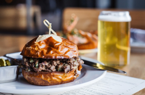 A close-up of a pretzel bun burger filled with ground meat, served with pickles and a glass of beer in the background.