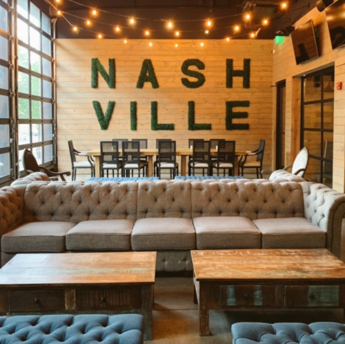 Cozy interior with a tufted sofa, wooden tables, and "NASHVILLE" greenery on the wall, illuminated by string lights.