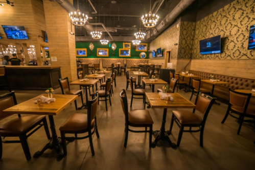A spacious restaurant interior with wooden tables, leather chairs, and decorative lighting, featuring multiple TVs on the walls.