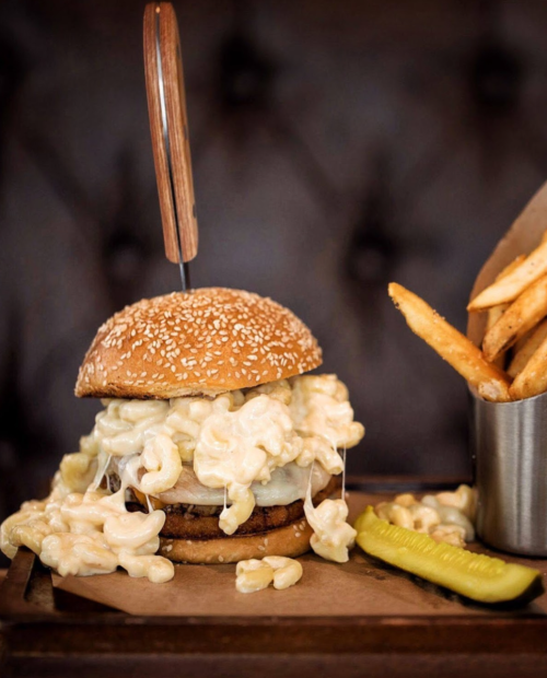 A towering burger topped with creamy macaroni and cheese, served with fries and a pickle on the side.