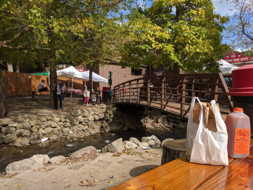 A scenic view of a bridge over a stream, with people nearby and food bags on a wooden table in the foreground.