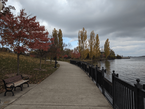 A scenic walkway by a lake, lined with trees in autumn colors and a bench, under a cloudy sky.