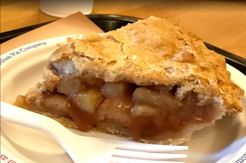 A slice of apple pie with a flaky crust, revealing a warm, gooey apple filling, served on a paper plate with a fork.