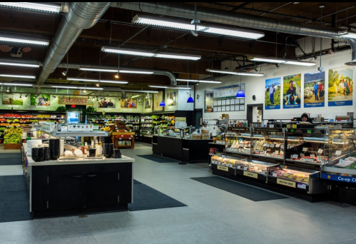 A grocery store interior featuring produce displays, a deli counter, and colorful wall murals.