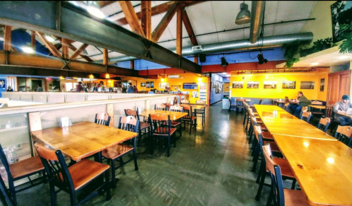 A spacious, brightly lit café with wooden tables, chairs, and a colorful interior, featuring exposed beams and a cozy atmosphere.