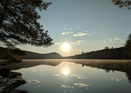 A serene lake at sunrise, with mist rising and mountains reflected in the calm water under a clear sky.