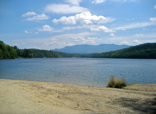 A serene lake surrounded by green hills and mountains under a blue sky with fluffy clouds.