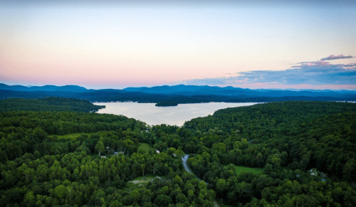 Aerial view of a serene lake surrounded by lush green forests and distant mountains under a pastel sky.