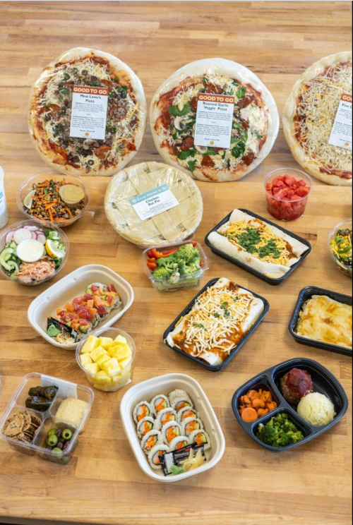 A variety of prepared meals and pizzas displayed on a wooden table, including salads, sushi, and fruit.